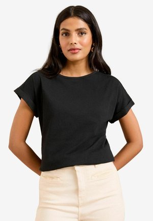 Pikkade tumedate juustega naine, kes kannab musta lühikese varrukaga särki ja helevärvilisi pükse, seisab käed selja taga.