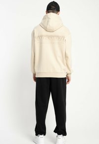 Mężczyzna w beżowej bluzie z kapturem z napisem "SANDS SHIFTER" na plecach, w czarnych spodniach, białych sneakersach i czapce baseballowej, odwrócony plecami na białym tle.