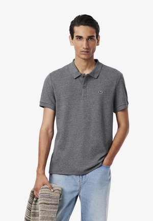 Polo de color gris oscuro con cuello abotonado, mangas cortas y un logotipo bordado en verde. El modelo sostiene un artículo de punto a rayas.