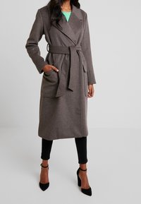 Brauner Wollmischung langer Mantel mit Wickeldesign, Bindegürtel, zwei vorderen Taschen und Reverskragen, getragen über schwarzen Hosen und hohen Schuhen.