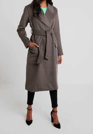 Brauner Wollmischung langer Mantel mit Wickeldesign, Bindegürtel, zwei vorderen Taschen und Reverskragen, getragen über schwarzen Hosen und hohen Schuhen.