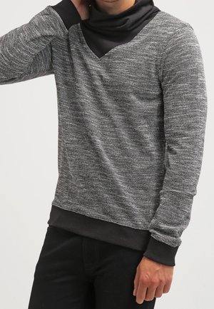 Homme portant un pull gris texturé à manches longues avec des poignets et un ourlet noirs, associé à un pantalon noir, main près du cou.