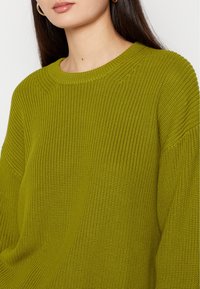 Grüner Strickpullover mit Rundhalsausschnitt, der eine strukturierte Oberfläche und lockere Ärmel hat. Der Stoff wirkt weich und bequem.