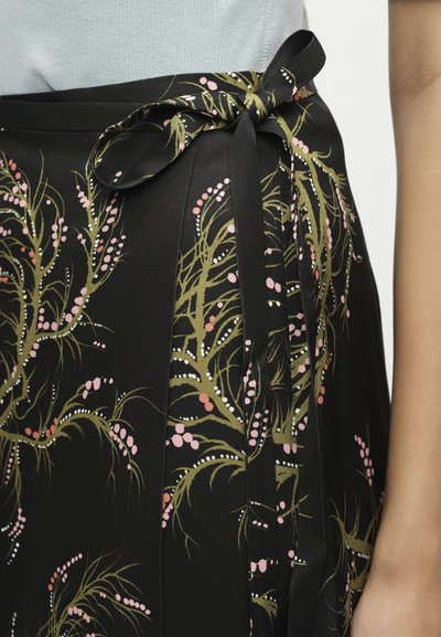 Μαύρη φούστα με πράσινο και ροζ λουλουδάτο σχέδιο, δεμένη στη μέση με φιόγκο, σε συνδυασμό με ανοιχτό γκρι τοπ, με το χέρι στη δεξιά πλευρά.