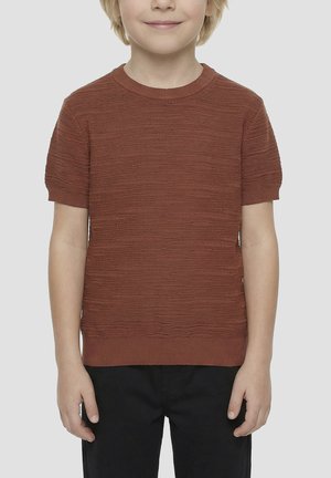 Enfant portant un pull texturé rouille à manches courtes et un pantalon noir, debout devant un fond clair uni.