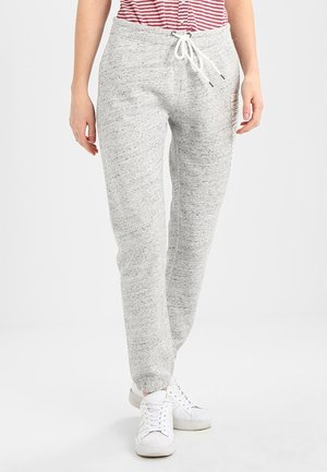 Henkilö, joka pukeutuu vaaleanharmaisiin collegehousuihin, joissa on nyörivyötärö ja joustavat resorit lahkeensuissa, sekä valkoiset lenkkarit, seisoo yksinkertaisen taustan edessä.