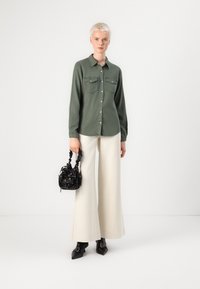Camisa de mangas longas verde-oliva com dois bolsos no peito, combinada com calças largas creme e botins pretos de bico fino. Segura uma bolsa preta.