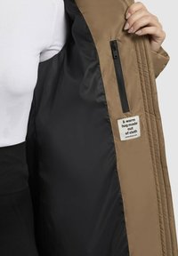 Pufová bunda s béžovým exteriérom a čierne podšívkou. Obsahuje zipsovú kapsu a nálepku s nápisom "Teplé objatie vyrobené z látky."