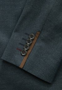 Mørkeblåt stof med et hørflet mønster; har fire sorte knapper med farvede tråde og en brun detalje på ærmet.