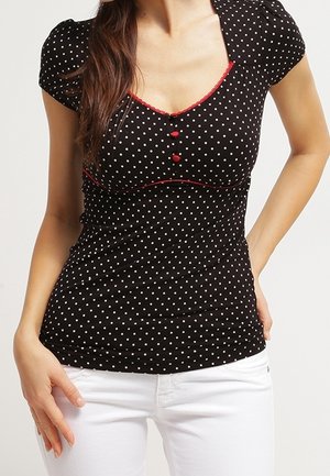 Vrouw draagt een zwart nauwsluitend topje met korte mouwen, witte polka dots, rode bies en knopen, gecombineerd met witte broek.