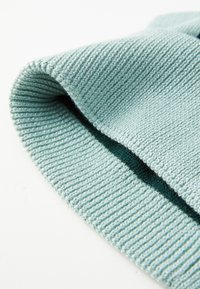 Tessuto lavorato a maglia di colore azzurro chiaro con una texture a coste, caratterizzato da un accento verde. I bordi sono arrotolati e rifiniti con cura, mostrando artigianalità.