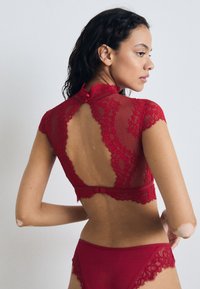 Červený čipkovaný bralet s vysokým golierom, krátkymi rukávmi a otvoreným dizajnom na chrbte. Zodpovedajúce červené čipkované nohavičky s kvetinovým vzorom a elastickým pásom.