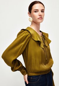 Olivenfarvet bluse med rynket halsudskæring og ærmer, samlet talje og snorelukning. Båret med mørke jeans og store cremede øreringe.