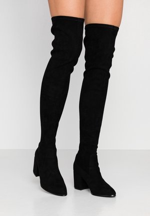 Personne portant des bottes en suède noires au-dessus du genou avec talons blocs, debout sur un sol blanc devant un fond uni.