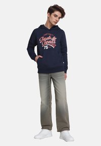 Tienerjongen staat met zijn hand in zijn zak, draagt een marineblauwe graphic hoodie, vervaagde grijze jeans en witte sneakers tegen een effen achtergrond.