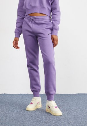 Person iført lilla Nike træningsbukser og matchende kort cropped sweatshirt, hvide sneakers med pink Nike logoer, står på gråt tæppe.