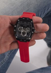 Black rectangular watch with a red silicone strap, featuring multiple dials and silver accents on a clear face, held in a hand.