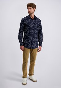 Mann trägt ein dunkelblaues Langarmhemd mit weißen Punkten, sandfarbene Hose und weiße Sneaker, steht vor einem schlichten hellen Hintergrund.