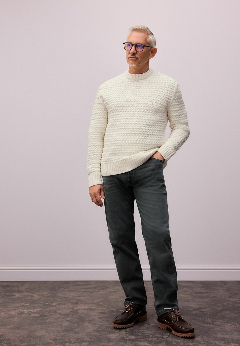 Cremefarbener, strukturierter Strickpullover mit geripptem Saum, graue Jeans und braune Schnürschuhe mit dicker Sohle; stehend vor einem neutralen Hintergrund.