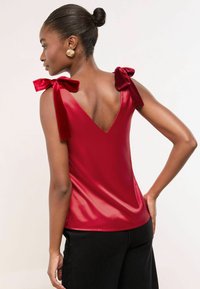 Piros selyem felső V-neccel és bársonyos masnikkal a vállakon, fekete nadrággal párosítva. Sima textúra és szakszerűen megvarrt fazon.