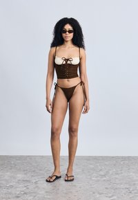Καφέ μαγιό δύο τεμαχίων με κορυφή σε στυλ κοντού τοπ που διαθέτει ρυθμιζόμενες τιράντες και λεπτομέρεια δέσιμου μπροστά, σε συνδυασμό με ταίριαστα σλιπ μπικίνι με κορδόνια.