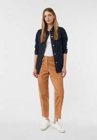 Camicia di jeans di colore blu scuro con bottoni frontali e due tasche sul petto, abbinata a pantaloni attillati color testa di moro e sneakers bianche. Tessuto dalla texture liscia.