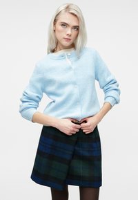 Mlada ženska z blond lasmi, ki nosi svetlo modri kardigan in temno zeleni ter modri karirani krilo, prilagaja pas svojega krila.