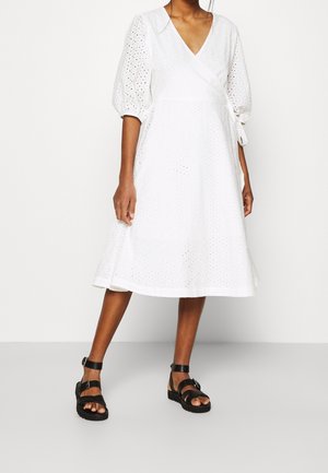 Weißes, knielanges Wickelkleid mit kurzen Puffärmeln, versehen mit Lochstickerei und einer Bindung in der Taille. Kombiniert mit schwarzen Riemchensandalen.