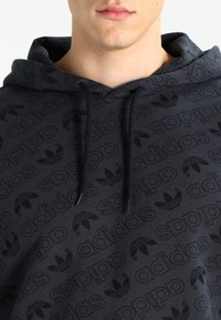 Muž v tmavé mikině s kapucí, na které se opakují loga Adidas a název značky, černými šňůrkami a viditelným výstřihem a rty.