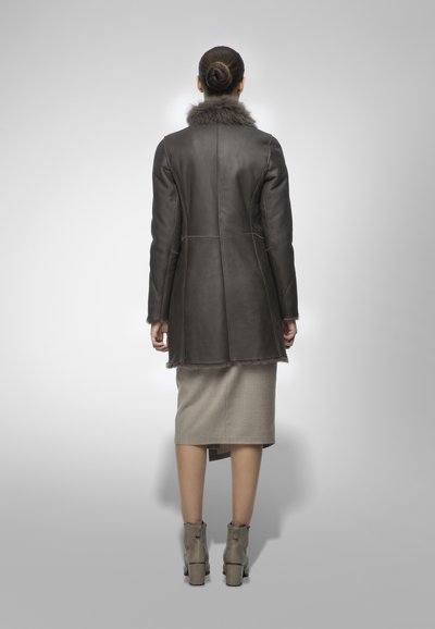 Cappotto in pelle marrone con colletto in pelliccia, vestibilità aderente, lunghezza media, caratterizzato da cuciture verticali. Indossato sopra una gonna a tubino grigia chiara e stivaletti alla caviglia.