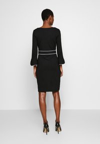 Abito nero aderente con maniche lunghe e polsini svasati. Presenta una cintura bianca a contrasto in vita. Texture liscia, design al ginocchio.