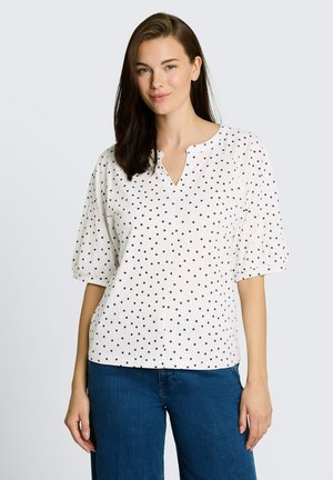 Femme aux cheveux foncés portant un chemisier blanc à pois noirs et un jean bleu, debout devant un fond gris clair.