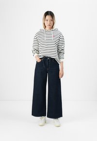 Femme se tenant debout avec les mains dans les poches, portant un hoodie rayé noir et blanc, un jean large sombre et des baskets blanches sur un fond blanc.