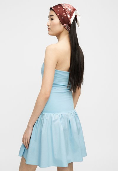 Νεαρή γυναίκα με μακριά μαύρα μαλλιά φοράει κόκκινο μαντήλι με σχέδια και αμάνικο ανοιχτό μπλε φόρεμα, στέκεται σε προφίλ μπροστά σε απλό φόντο.