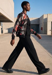 Frau geht draußen in schwarzen Hosen, schwarzem Oberteil und einer schwarzen Blumenjacke vor einem modernen Betongebäude und klarem blauem Himmel.