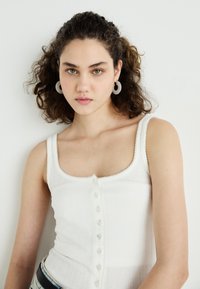 Top in cotone bianco a coste con scollatura a girocollo, chiusura decorativa a bottoni e bordo a onde. A vista un paio di orecchini a cerchio texturizzati.