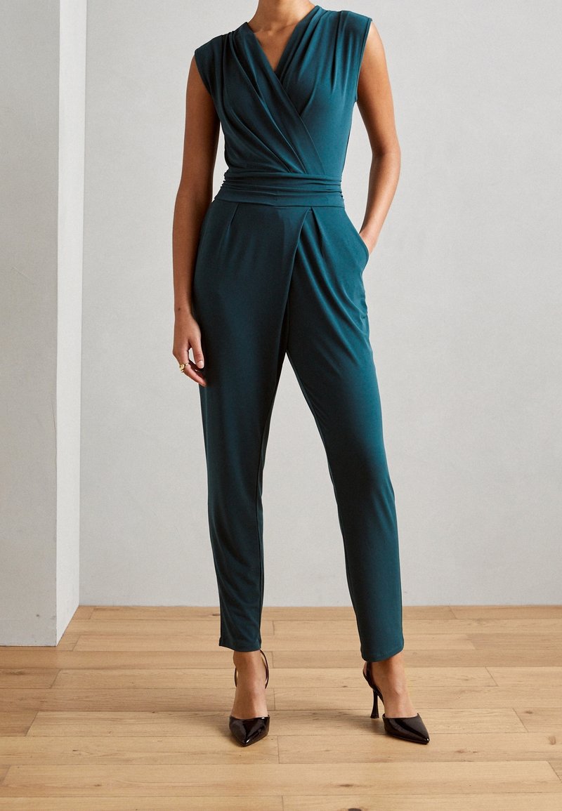 Mujer vestida con un mono sin mangas en color teal, con un escote envolvente, pantalones ajustados y tacones altos de punta negra, de pie sobre un suelo de madera.