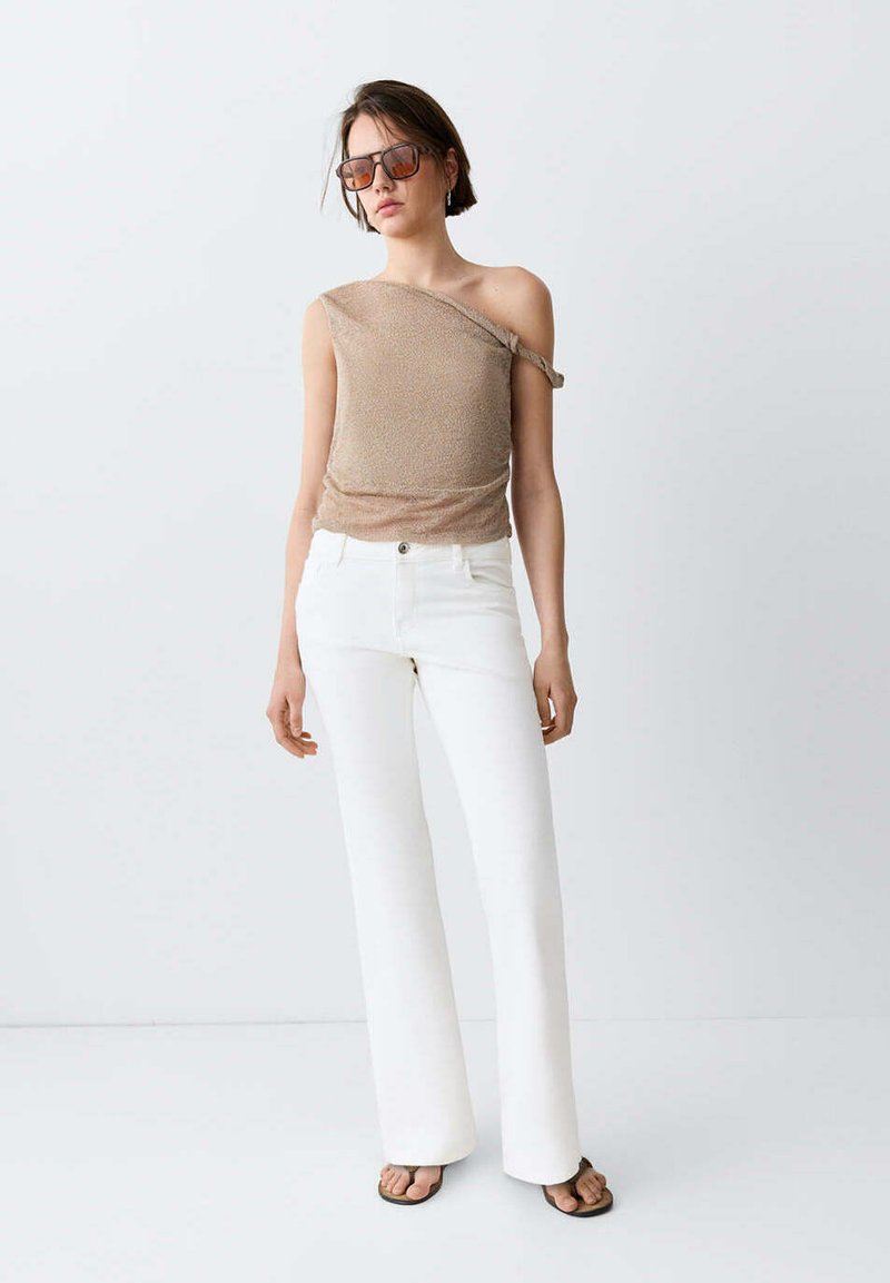 Femme portant un top marron asymétrique, un pantalon blanc taille haute à jambes larges, des lunettes de soleil et des sandales noires, debout devant un fond blanc.
