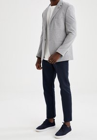 Hellgrauer Blazer mit einem Knopf und einer Brusttasche, kombiniert mit einem weißen T-Shirt, marineblauen Hosen und dunklen Sneakers.