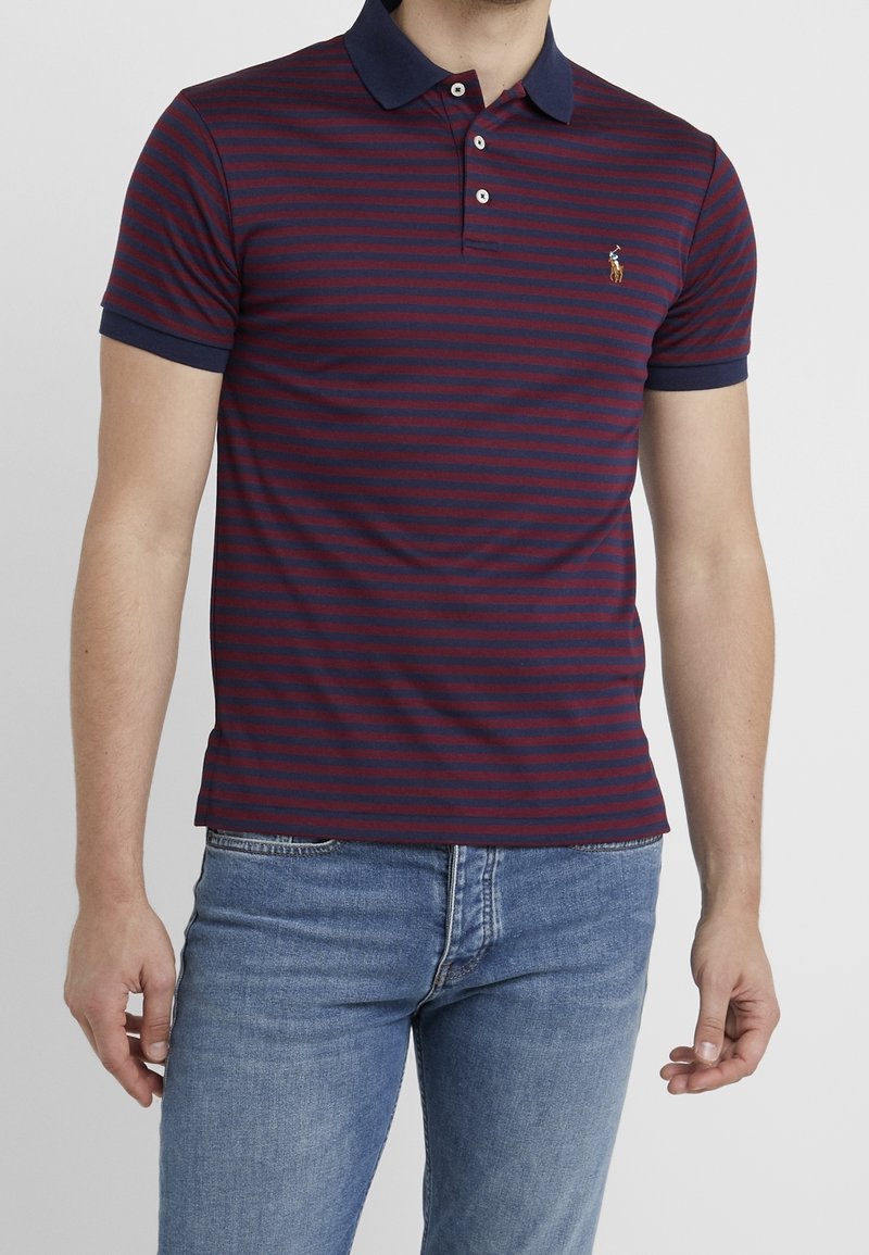 Homme portant un polo à manches courtes rayé bleu marine et bordeaux avec un logo brodé et un jean bleu clair, debout devant un fond gris uni.