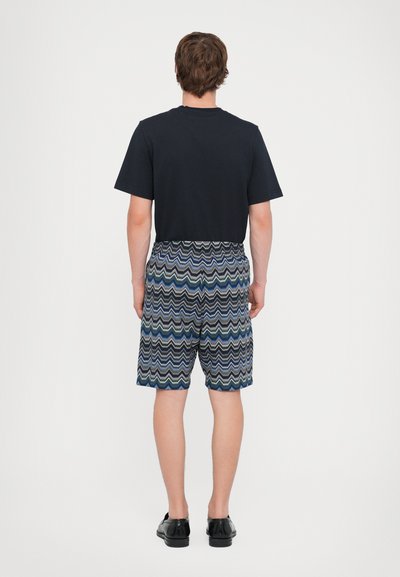 Mörkblå T-shirt, bakvy, ihopparad med mönstrade shorts som har vågiga linjer i blått och grönt på en mörk bakgrund. Svarta skor fulländar looken.