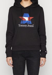 Person som bär en svart hoodie med en design av blå, vit och röd stjärna samt texten "Tommy Jeans" på framsidan, kombinerad med svarta byxor.