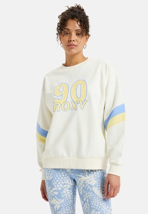 Light beige sweatshirt with blue and yellow stripes on sleeves, featuring "90" and "ROXY" in bold lettering, made from soft fabric.