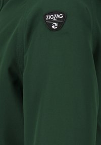 Groene jas van gladde stof met een zwart driehoekig logo-appliqué met de tekst "ZIGZAG" op de linker bovenkant.