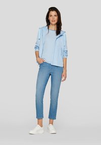Helle blaue leichte Jacke über einem weiß-blau gestreiften Hemd, kombiniert mit hellblauen Jeans und weißen Sneakers. Einfaches Design und lässige Passform.