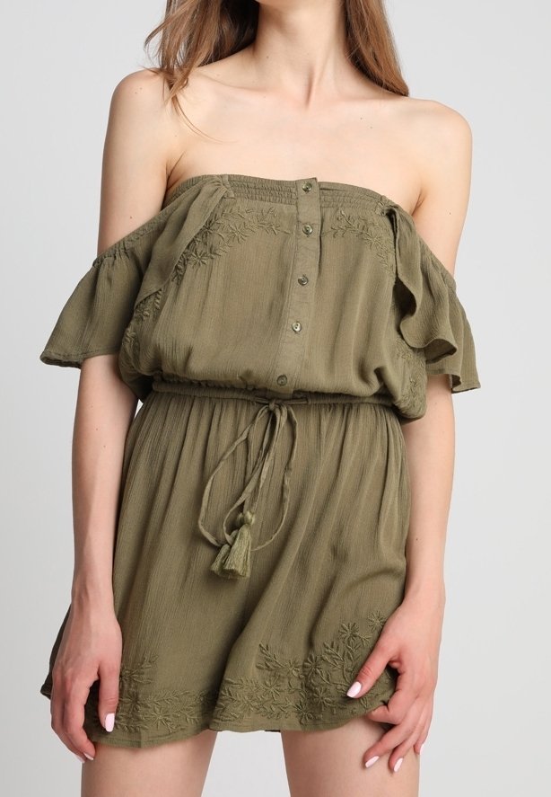 Femme portant une combishort vert olive épaules dénudées avec boutons, détails floraux brodés et ceinture à franges à la taille.