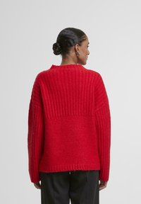 Czerwony sweter z dzianiny o fakturowanym prążkowanym wzorze, z długimi rękawami i okrągłym dekoltem, wykonany z miękkiej wełnianej tkaniny.