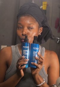 Två 300 ml blå plastflaskor med Redken hårprodukter, märkta "extreme anti-snap" och "extreme stay 220," hålls i händerna.