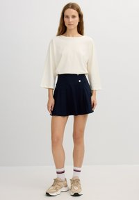 Falda plisada navy con cintura alta y un detalle de botón lateral, combinada con una blusa de mangas largas color crema claro y zapatillas beige.