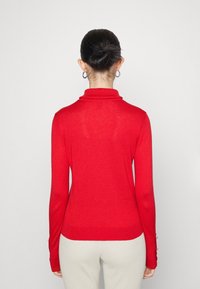 Punainen, pitkähihaine turtleneck-neule, jossa on ribbitekstuuri, tiukka muotoilu ja nappikoristeet hihansuissa.